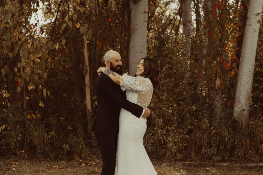 A couple is standing in front of some trees, hugging each other and laughing on their elopement day.